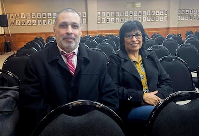 Liliana González de Bristot, con chaqueta negra y blusa amarilla, y Mario Elizeche, con abrigo negro y corbata de rayas, conversan en un auditorio vacío.
