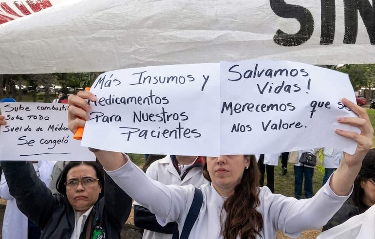 Grupo de médicos con batas blancas sosteniendo carteles de protesta sobre insumos y valorización del trabajo en un entorno al aire libre.