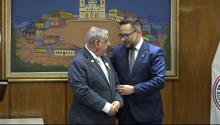Derlis León en traje azul oscuro y corbata gris, conversando con Isaías Ricardo Fretes, en un ambiente gubernamental elegante.