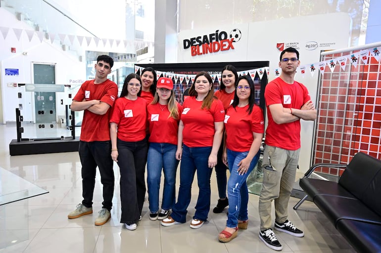 Nueve personas con camisetas rojas posan en un ambiente moderno, sonriendo y mostrando felicidad en el evento 'Desafío Blindex'.