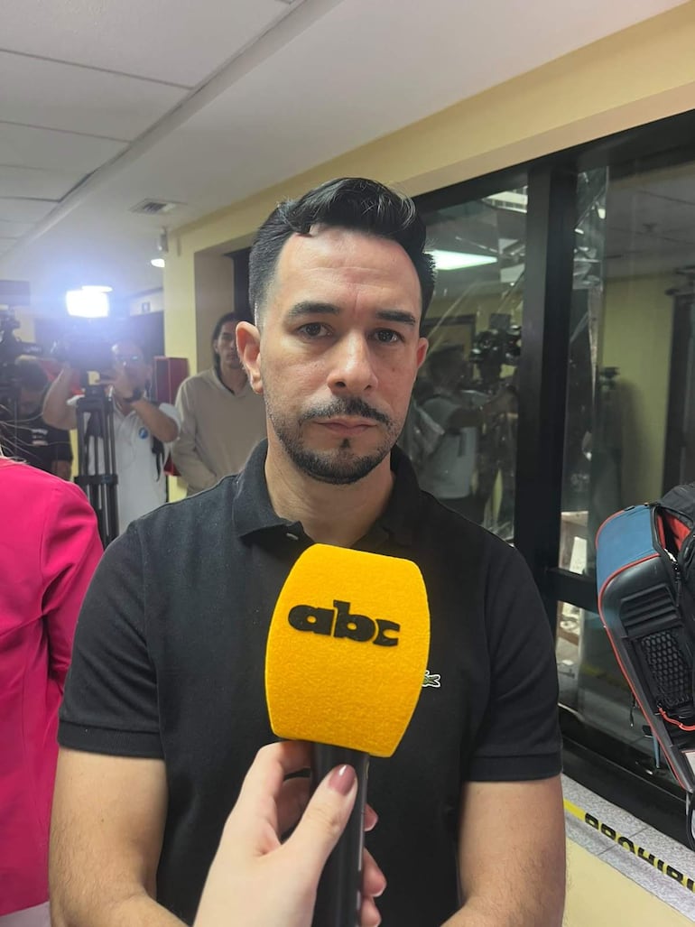 Sebastián Romero, con camiseta negra y barba, sostiene un micrófono mientras una mujer con blusa rosa lo escucha atentamente.