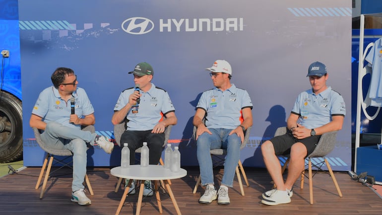 Cuatro hombres en camisetas azules de Hyundai, hablando y escuchando en un ambiente al aire libre con logo del equipo de fondo.