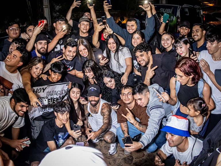 Arcángel con camiseta blanca y tatuajes, rodeado de jóvenes sonriendo y tomando selfies en un evento al aire libre.