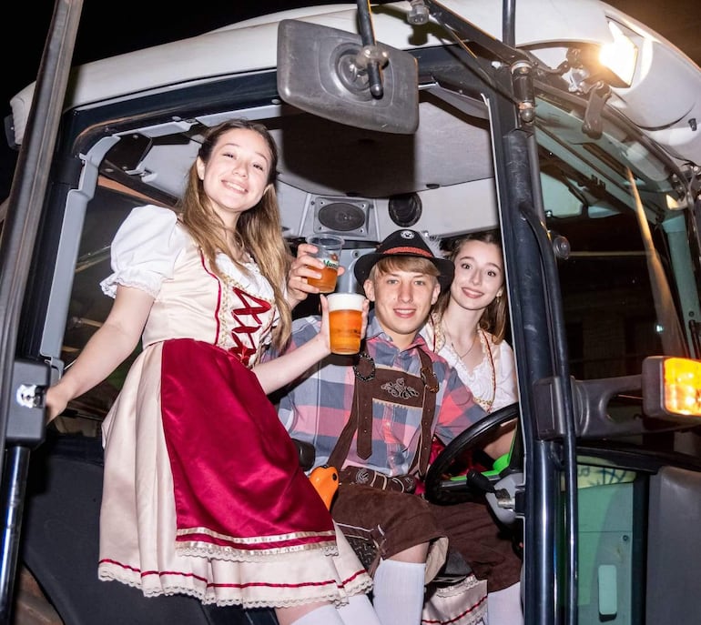 Mujer con vestido tradicional sostiene vaso, sonríe junto a un hombre con camisa a cuadros y otra mujer similar en un ambiente festivo.