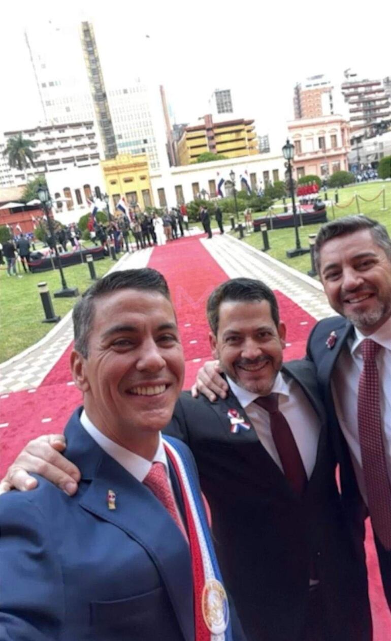 Santiago Peña, Raúl Latorre y Pedro Alliana sonríen para una selfie, vestidos formalmente, en un entorno festivo con banderas.