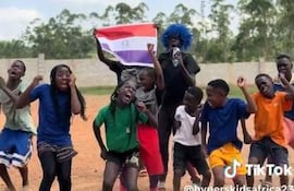 Grupo de niños y adolescentes en camiseta colorida, uno sosteniendo una bandera paraguaya, bailando al aire libre con expresiones alegres.