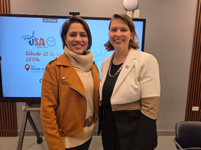 Dos mujeres sonrientes, una con abrigo marrón y la otra en atuendo blanco, posan en un entorno moderno frente a una pantalla del evento.
