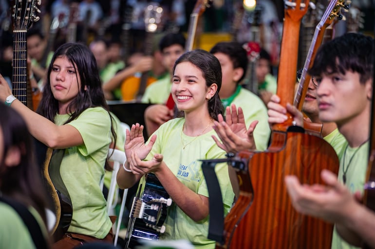 Chicas sonrientes, una con guitarra, otra aplaudiendo, rodeadas de jóvenes con camisetas verde claro en ambiente musical.