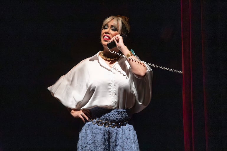 Mujer de pie en el escenario, con blusa blanca y falda azul, sosteniendo un teléfono mientras actúa en un fondo oscuro.