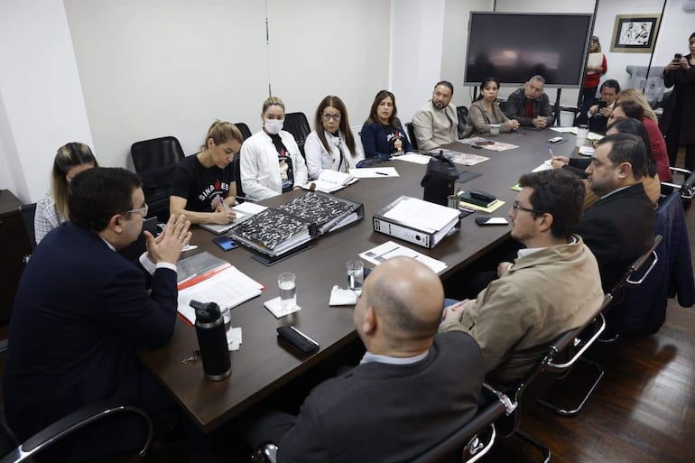 Hombre en traje conversando, mujer con camiseta de Sinamed tomando notas, otros participantes observan atentamente en una sala de conferencia.