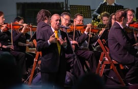 Un hombre en traje negro y corbata amarilla actúa frente a un micrófono, rodeado de músicos de orquesta con instrumentos de cuerda.