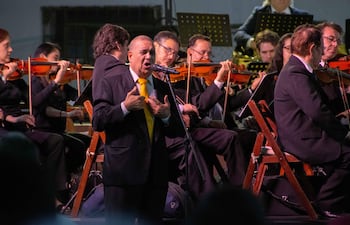 Un hombre en traje negro y corbata amarilla actúa frente a un micrófono, rodeado de músicos de orquesta con instrumentos de cuerda.