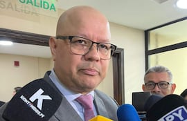 Hombre calvo y con gafas en traje gris habla a la prensa, rodeado de micrófonos en un ambiente iluminado.