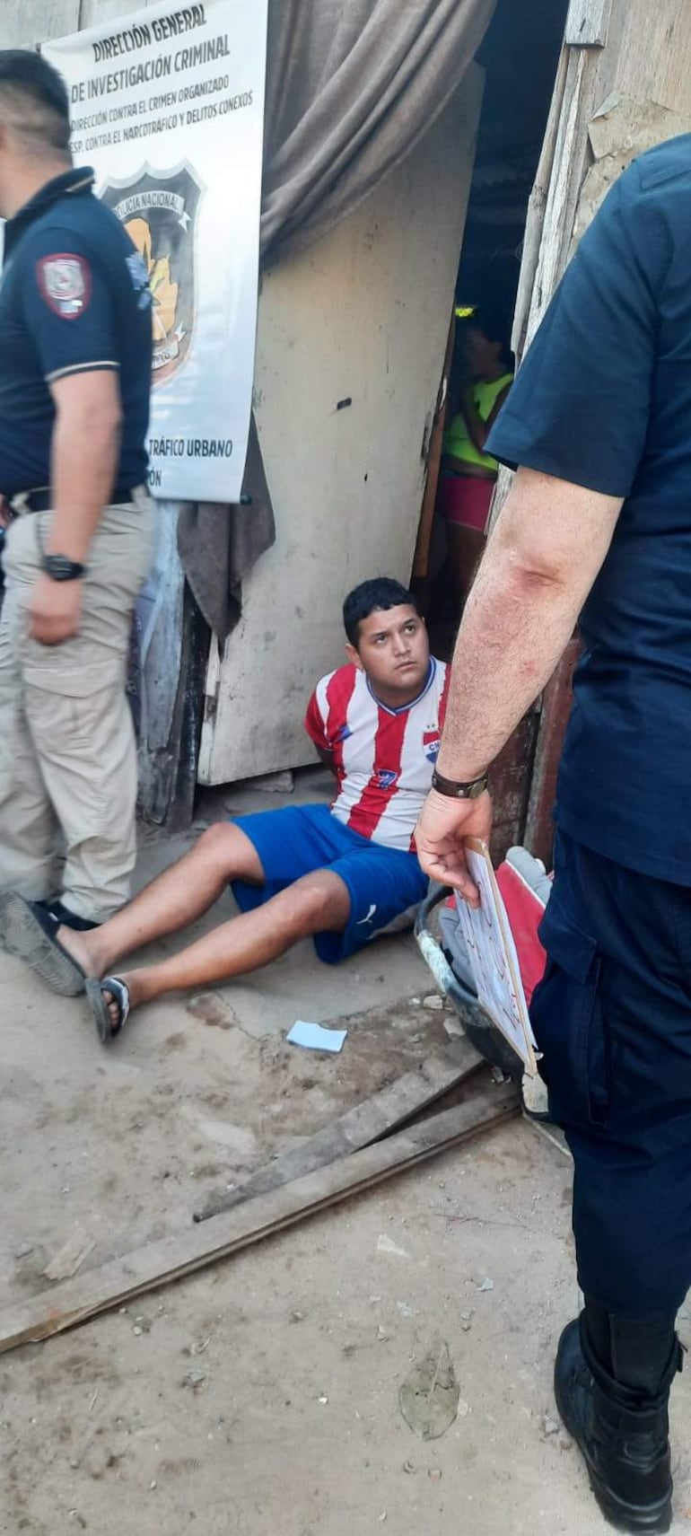 Joven con camiseta de rayas de Club Olimpia, sentado en el suelo, rodeado por varios policías con documentos en mano.