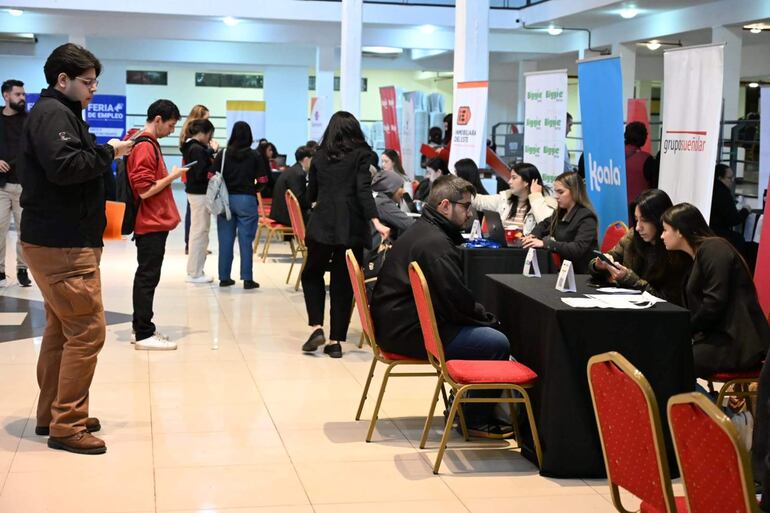Tres jóvenes en mesa de feria de empleo, uno conversando y dos revisando sus teléfonos en un ambiente moderno y dinámico.