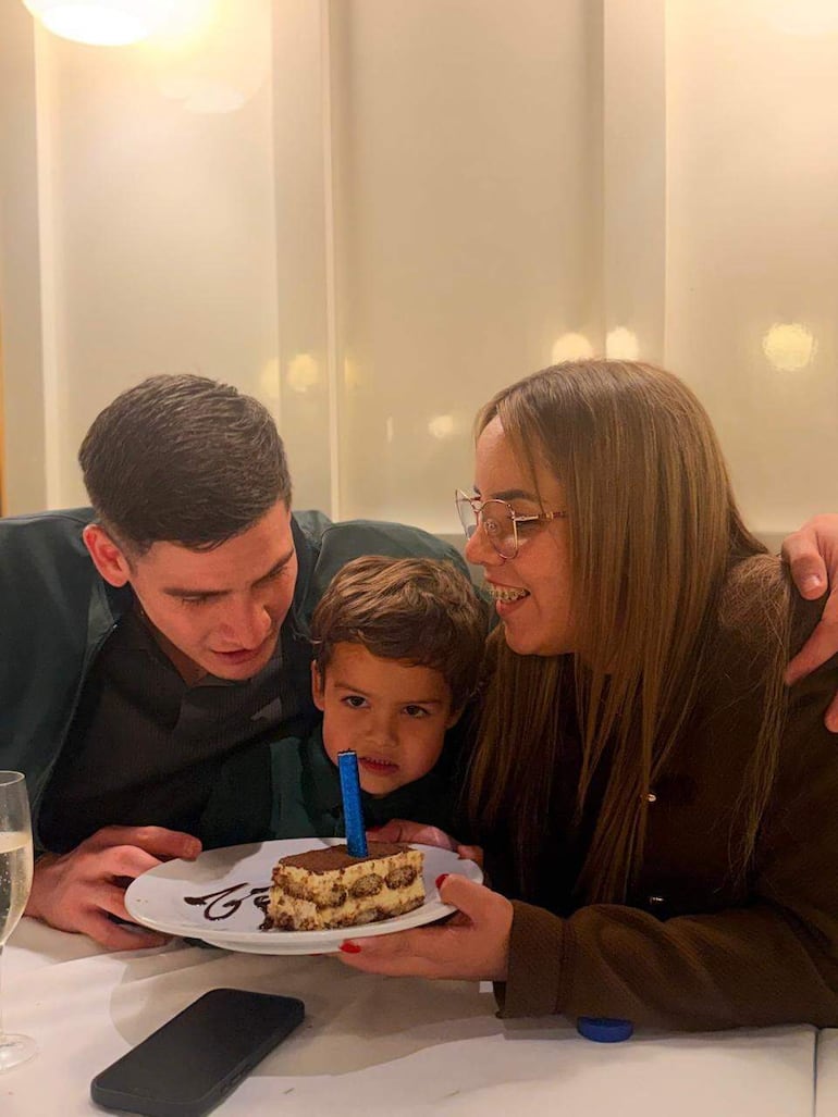 Niño sosteniendo un pastel con vela azul. Hombre sonriente a la izquierda y mujer a la derecha, todo en un ambiente acogedor.