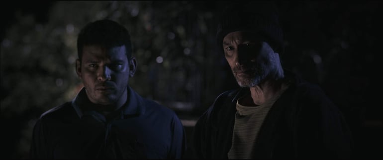 Dos hombres, uno con camiseta azul y otro en camiseta clara y gorro negro, conversan en un ambiente oscuro y tenso.