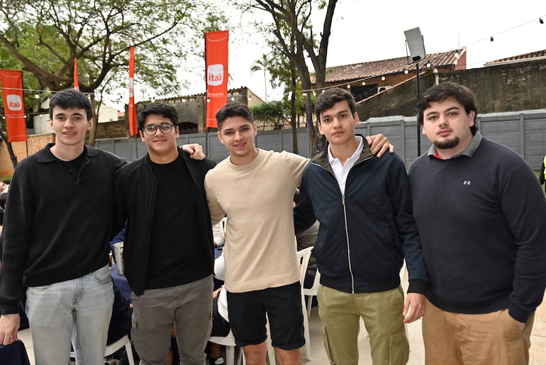 Cinco jóvenes de pie, sonriendo, con ropa casual y brazos sobre los hombros en un ambiente festivo con banderas de Itaú.
