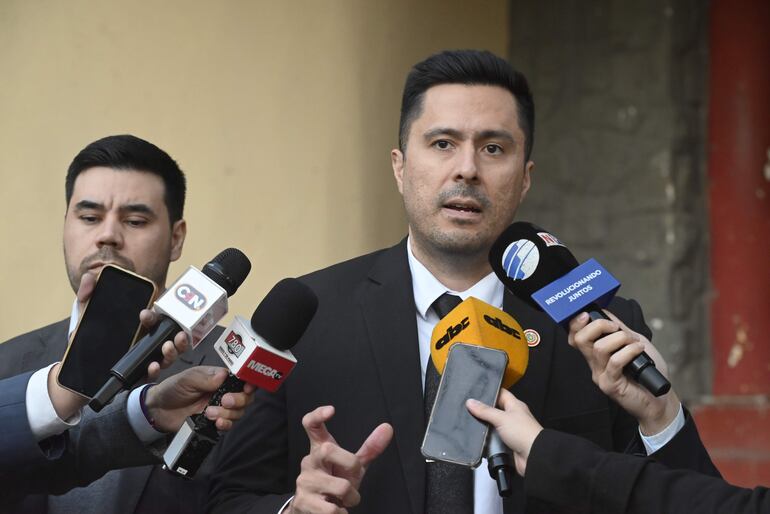 Hombre en traje oscuro habla con micrófono, rodeado de periodistas con grabadoras y un teléfono móvil en mano.