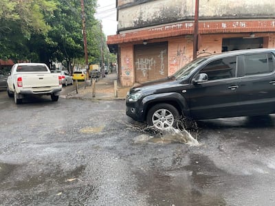 Un vehículo negro salpica agua mientras gira en una calle con baches, rodeado de otros autos y un edificio deteriorado.