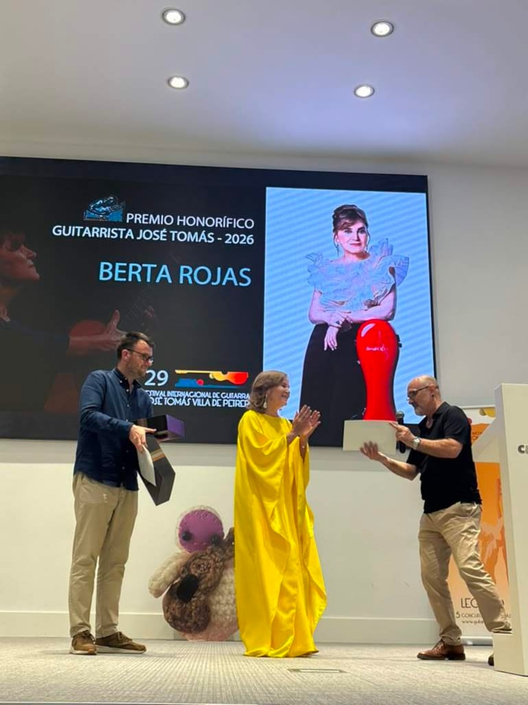 Berta Rojas con capa amarilla, sonriente, recibe un premio mientras dos hombres presentan documentos en un ambiente formal.