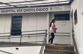 Mujer con camiseta a rayas rojas y blancas camina hacia la recepción del hospital, rodeada de un entorno arquitectónico sencillo.