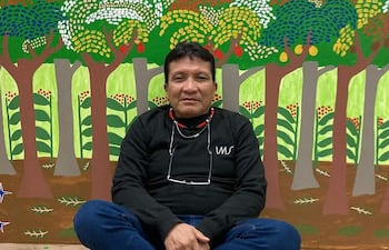 Joseca Yanomami, hombre con camiseta negra y jeans azules, sentado en el suelo frente a un mural colorido de paisaje natural.