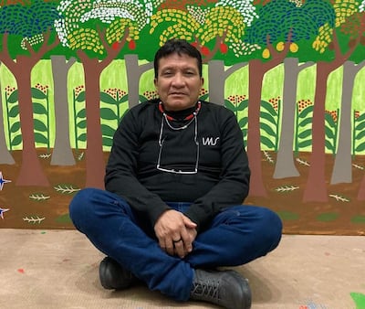 Joseca Yanomami, hombre con camiseta negra y jeans azules, sentado en el suelo frente a un mural colorido de paisaje natural.
