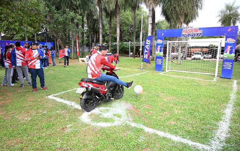Hombre con camiseta roja y rayas azules patea un balón, rodeado de público en un parque con árboles y césped.