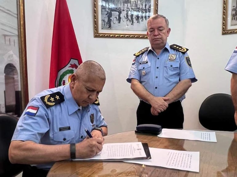 Juan Ramón Agüero Ríos, vestido de uniforme policial, firma un documento mientras César Silguero lo observa con expresión seria.
