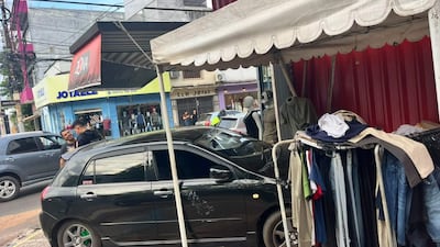 Dos hombres conversan en la calle, uno sonríe y lleva camiseta negra, con edificios y un local comercial de fondo.