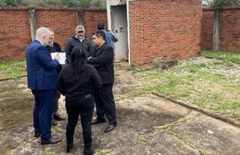 Héctor Daniel Granada en traje azul sostiene documentos, mientras otros participan en la conversación, rodeados de un área verde descuidada.