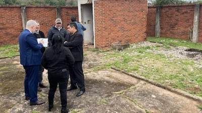 Héctor Daniel Granada en traje azul sostiene documentos, mientras otros participan en la conversación, rodeados de un área verde descuidada.