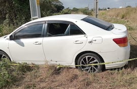 Automóvil blanco dañado con orificios y ventanas rotas, estacionado en una zona semiurbana con pasto seco alrededor.