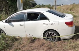 Automóvil blanco dañado con orificios y ventanas rotas, estacionado en una zona semiurbana con pasto seco alrededor.