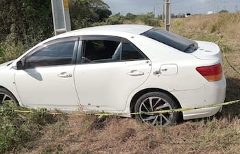 Automóvil blanco dañado con orificios y ventanas rotas, estacionado en una zona semiurbana con pasto seco alrededor.
