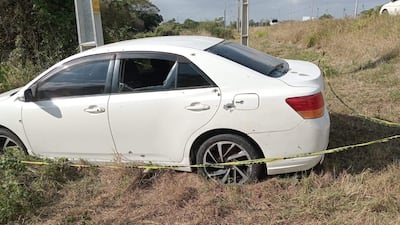 Automóvil blanco dañado con orificios y ventanas rotas, estacionado en una zona semiurbana con pasto seco alrededor.