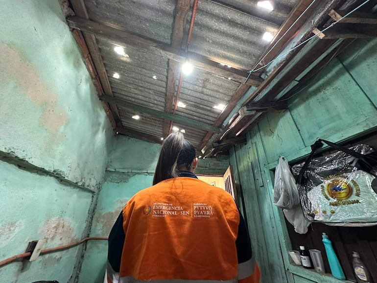 Funcionaria con chaqueta de emergencia naranja y negra observa una vivienda dañada, en un ambiente con paredes verdes.
