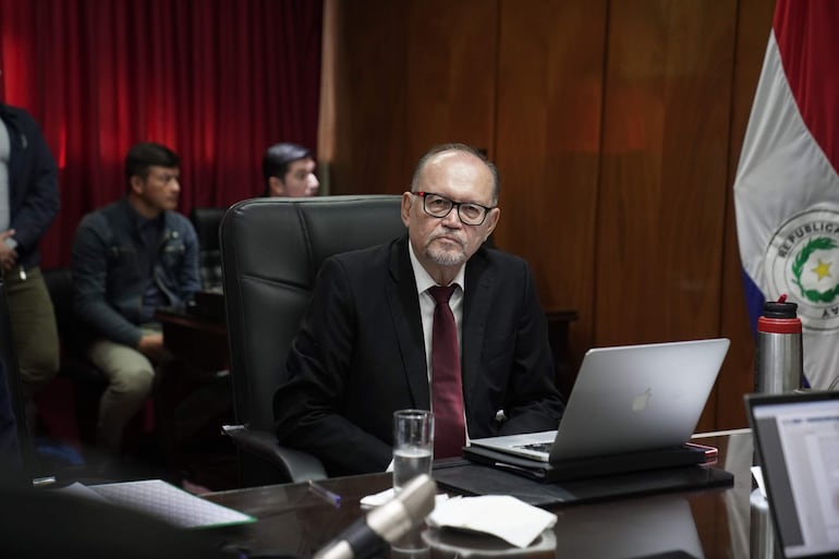 Hombre canoso con gafas en traje negro sentado en mesa, junto a asistentes con atuendos informales y formales, en ambiente de oficina.