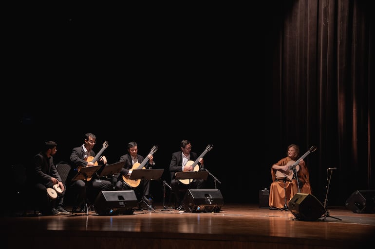 Berta Rojas con guitarra en prenda marrón, rodeada de músicos concentrados en un escenario con luz tenue.