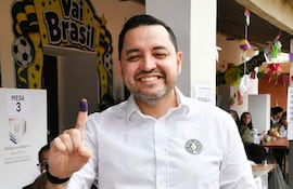 Luis Bello sonriente con camisa blanca y dedo índice teñido de morado en un centro de votación decorado.
