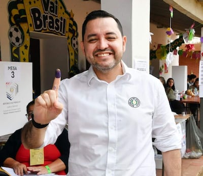 Luis Bello sonriente con camisa blanca y dedo índice teñido de morado en un centro de votación decorado.