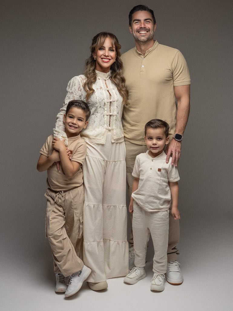 Familia compuesta por madre con blusa blanca, padre en camiseta beige, y dos niños con ropa clara, todos sonrientes en un fondo neutro.