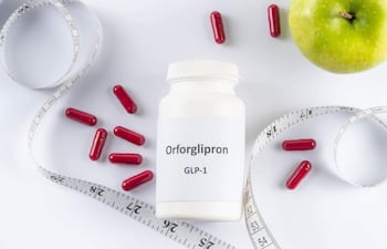 Frasco blanco de Orforglipron con cápsulas rojas sobre superficie blanca, rodeado de una manzana verde y cinta métrica.