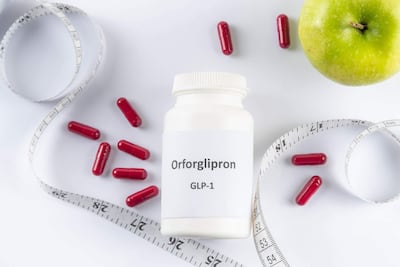 Frasco blanco de Orforglipron con cápsulas rojas sobre superficie blanca, rodeado de una manzana verde y cinta métrica.