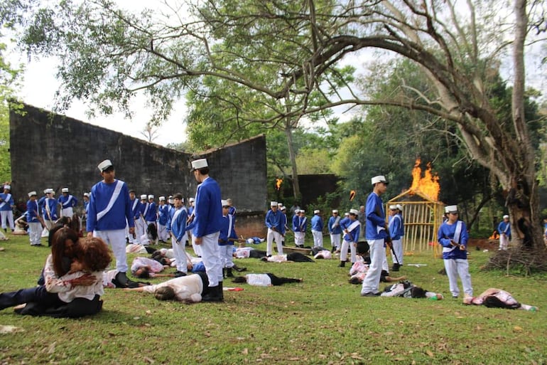 Jóvenes en uniformes azules y blancos, algunos agachados y otros de pie, en un campo con una estructura en llamas al fondo.
