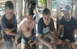 Cinco hombres con expresiones serias, uno con herida en la cabeza, sentados en un entorno rural entre árboles y mesas.