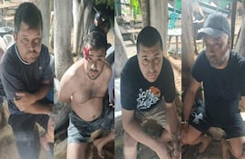 Cinco hombres con expresiones serias, uno con herida en la cabeza, sentados en un entorno rural entre árboles y mesas.