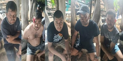 Cinco hombres con expresiones serias, uno con herida en la cabeza, sentados en un entorno rural entre árboles y mesas.