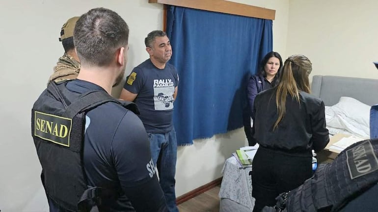 Herman Cecilio Melgarejo Morales escucha en el centro, rodeado de oficiales de SENAD y otros civiles en una habitación con cortinas azules.